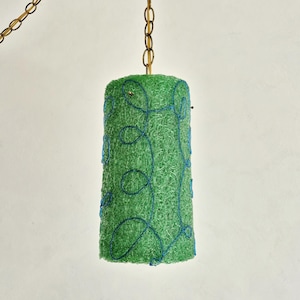 Peut inclure: Suspension luminaire cylindrique vert avec des coutures décoratives bleues. La lampe est suspendue à une chaîne dorée. L'abat-jour a un aspect tissé texturé, créant un élément d'éclairage unique.