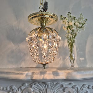 May include: A small, ornate chandelier with a gold-coloured metal frame and a globe of clear crystals. The light fixture hangs from a gold chain. A vase of white flowers sits nearby.