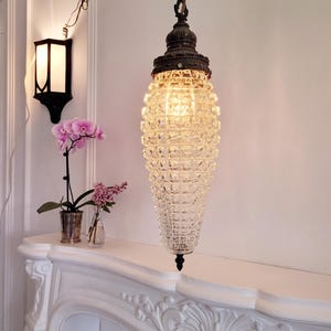 May include: A vintage glass and metal pendant light fixture with a clear, faceted glass shade hanging from a chain. The light is on and illuminating the room. The light fixture is hanging above a white fireplace mantel.