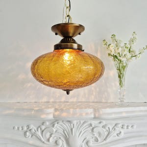 May include: A vintage amber glass pendant light. The textured, oval-shaped glass shade hangs from a brass-toned metal fixture. The light is on, emitting a warm glow. The fixture is suspended from a chain, suitable for a living room or dining room.