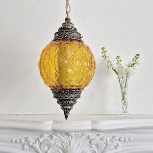 Puede incluir: Una lámpara colgante vintage de cristal ámbar con detalles metálicos ornamentados. La lámpara tiene una pantalla con forma de globo texturizado y cuelga de una cadena. La metalistería decorativa es oscura y detallada. La lámpara está suspendida sobre una repisa blanca.