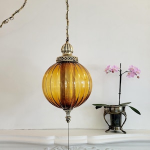以下が含まれることがあります： 装飾的な金属のディテールが施された、ヴィンテージの琥珀色のガラスペンダントライト。地球儀型の照明器具はチェーンから吊り下げられ、垂直のリブのディテールが特徴です。プルチェーンが下からぶら下がっています。近くには小さな蘭の植物があります。