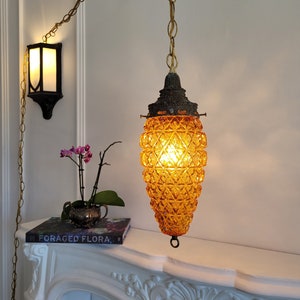 May include: A vintage amber glass pendant light with a geometric pattern hangs from a gold chain. The light is on and illuminating the room. The light is hanging above a white mantelpiece with a book titled "Foraged Flora" on top.