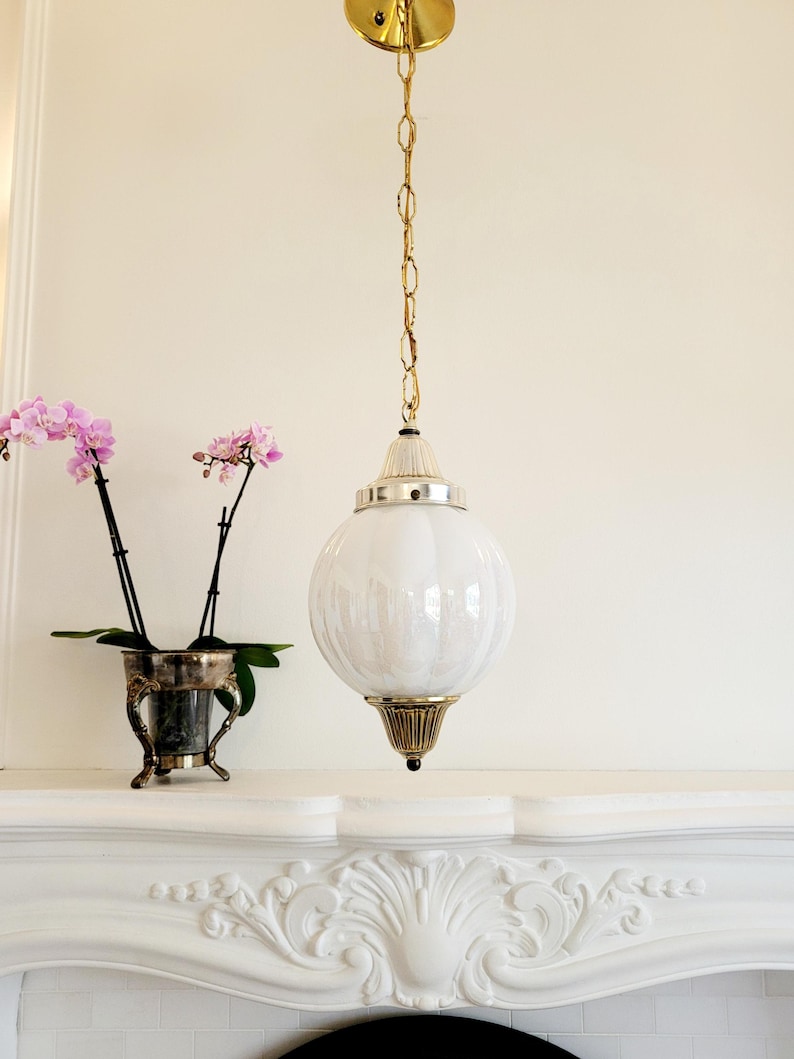 May include: A white globe pendant light fixture with a gold chain and a gold ceiling mount. The light fixture is hanging above a white fireplace mantel.