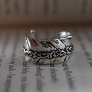 May include: A silver ring featuring a leaf design with intricate detailing. The ring is open and has a textured surface.