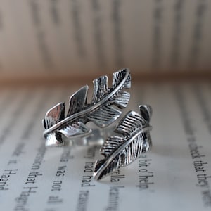 Puede incluir: Un anillo plateado con forma de dos plumas estilizadas, sobre un libro abierto. El diseño del anillo presenta detalles intrincados, con cada pluma con una superficie texturizada. El anillo es de estilo envolvente ajustable.