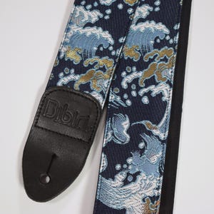 Peut inclure: Une sangle de guitare bleu marine avec un motif de vagues répétées en bleu clair, blanc et or. La sangle a une pièce d'extrémité en cuir noir avec le nom de la marque "Bibibi" en relief. Conçue pour les guitares.