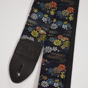 Puede incluir: Una correa de guitarra con un estampado floral azul oscuro con flores amarillas, naranjas y blancas. La correa tiene un extremo de cuero negro con el nombre de la marca "Bibiri" en relieve. La correa está diseñada para una guitarra.