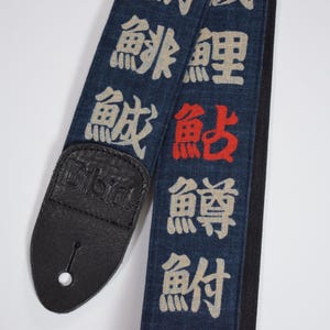 May include: A denim guitar strap with Japanese characters in white and red. The strap has a black leather end with the brand name "Dibin" embossed. The strap is designed for musical instruments.