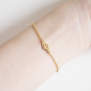 Puede incluir: Un brazalete de cadena dorada con un colgante geométrico. El colgante es un diseño hueco y abierto con forma de diamante.