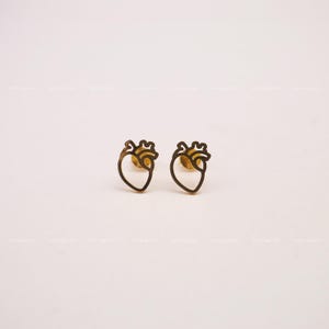 Puede incluir: Un par de pendientes de botón de corazón de color dorado. Los pendientes están hechos de un metal fino y tienen un diseño simple y minimalista.