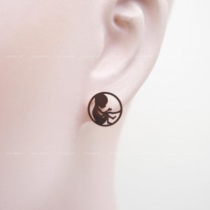 May include: A pair of round, dark brown stud earrings. Each earring features a cutout silhouette of a fetus within a circular frame. The earrings are displayed on a person's ear.