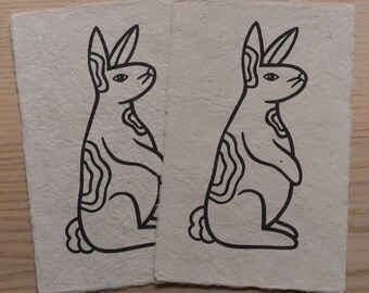 May include: Two pieces of handmade paper with a black line drawing of a rabbit. The rabbit is in profile, with long ears and a stylized body. The paper has a rough, textured surface and a natural, off-white color.