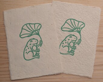 May include: Two rectangular, off-white paper cards with a green illustration of a frog holding a leaf. The frog has a dotted pattern and is under the leaf. The cards are on a light wood surface.