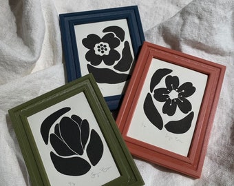 May include: Three framed black and white floral prints with a white background. The frames are green, blue, and red. The prints are signed "Sugi Boon" and include the size "1/2x".