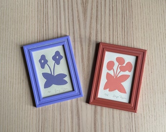 May include: Two framed prints of floral designs. The print on the left is purple and the print on the right is orange. Both prints have the text "#16 Song Poem" at the bottom.