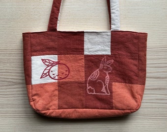 May include: A red and white checkered tote bag with a printed image of an orange and a rabbit. The bag has a brown fabric handle.