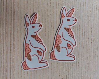 May include: Two white rabbit stickers with floral designs. The rabbits are facing to the right and have pink and orange flowers on their bodies.