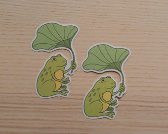 May include: Two stickers featuring cartoon frogs holding green lily pads. The frogs are light green with yellow bellies and are set against a light wood grain background. The stickers have a white border.