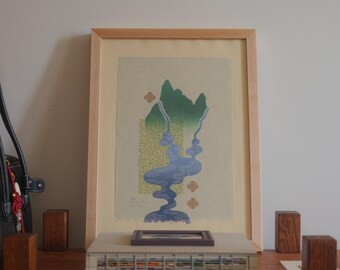 May include: Framed art print featuring a mountain landscape with a river in shades of blue and green. The artwork is in a light wood frame and rests on a wooden surface. Decorative wooden blocks are placed around the artwork.