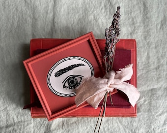 May include: A red framed picture with a black and white illustration of an eye with an eyebrow. The picture is sitting on top of two red books with a bundle of dried lavender flowers and a piece of beige fabric tied around them.