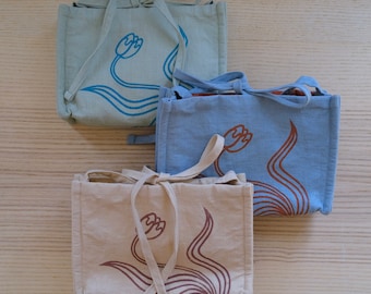 May include: Three small tote bags in light blue, blue, and beige, each featuring a stylized floral design in a contrasting color. The bags have matching tie closures and handles. The bags are made of a textured fabric.