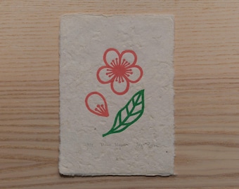 May include: A rectangular piece of textured paper with a red flower, a petal, and a green leaf design. The artwork is hand-printed with the text "Pink Blossom" and other handwritten text at the bottom.