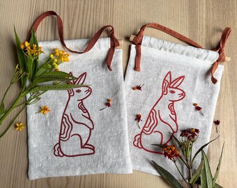 May include: Two white linen tote bags with red bunny rabbit illustrations. The bags have brown straps and are hanging on a wooden surface.
