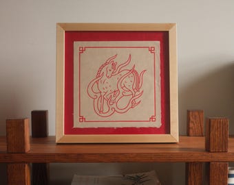 May include: Framed artwork featuring a red line drawing of a horse-like creature on a beige background, set against a red mat. The artwork is enclosed in a light wood frame. The piece is displayed on a wooden shelf with small wooden blocks.