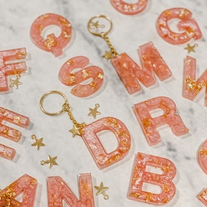 May include: Pink resin letter charms with gold flecks and star charms attached to a gold chain. The letters are A, B, C, D, F, G, M, N, Q, R, and S.