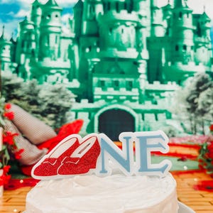 Peut inclure: Un gâteau blanc glacé avec un décor de chaussures rouges à paillettes et un décor de gâteau "ONE" bleu. L'arrière-plan présente un château vert et un ciel bleu, créant une scène de fête fantaisiste.