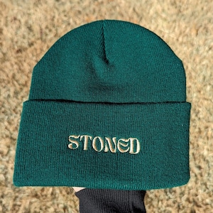 May include: A dark green beanie with the word "STONED" embroidered in gold on the front.