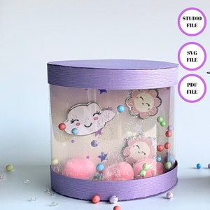 May include: A purple cylinder box with a clear window showing a whimsical scene with a cloud, a flower, and colorful pom-poms. The box is decorated with glitter and stars. The text "STUDIO FILE", "SVG FILE", and "PDF FILE" is visible in the image.