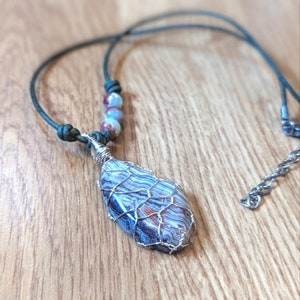May include: A silver wire wrapped pendant necklace featuring a blue and brown agate stone. The necklace has a green leather cord and silver beads.