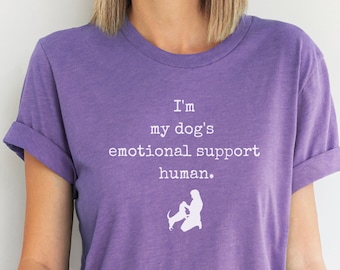 May include: Purple t-shirt with white text that reads "I'm my dog's emotional support human."  A white silhouette of a person kneeling with a dog is below the text.