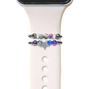 May include: Two beaded bracelets with a silver star charm, designed to fit around a white Apple Watch band. The beads are a mix of purple, blue, and gray.