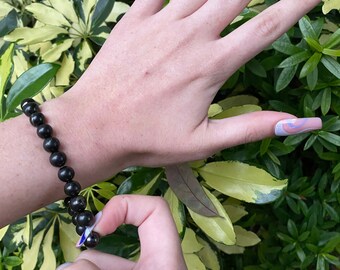 May include: A black beaded bracelet with round beads. The bracelet is on a person's wrist. The person's hand is holding the bracelet to show the beads.