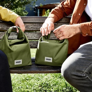 May include: Two people sitting on a wooden bench with green canvas bags. The bags have a simple design and a small label on the front.