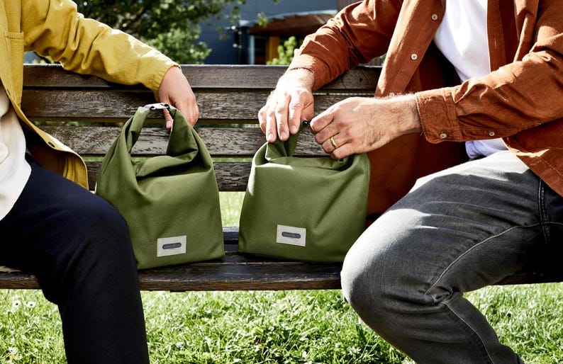 May include: Two people sitting on a wooden bench with green canvas bags. The bags have a simple design and a small label on the front.