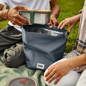 May include: A blue insulated lunch bag with a silver lining. A stainless steel lunch container with a wooden lid is being placed inside the bag.