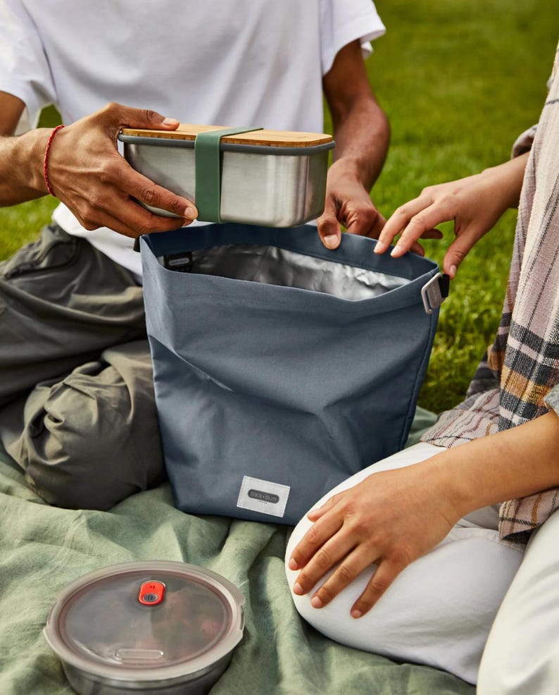 May include: A blue insulated lunch bag with a silver lining. A stainless steel lunch container with a wooden lid is being placed inside the bag.
