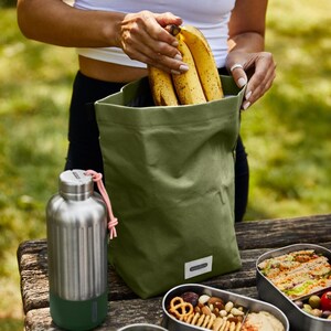 May include: A green canvas lunch bag with a pink drawstring, filled with bananas.  A stainless steel water bottle with a green bottom and a silver top is next to the bag.  Three stainless steel lunch containers are on a wooden table, filled with food.