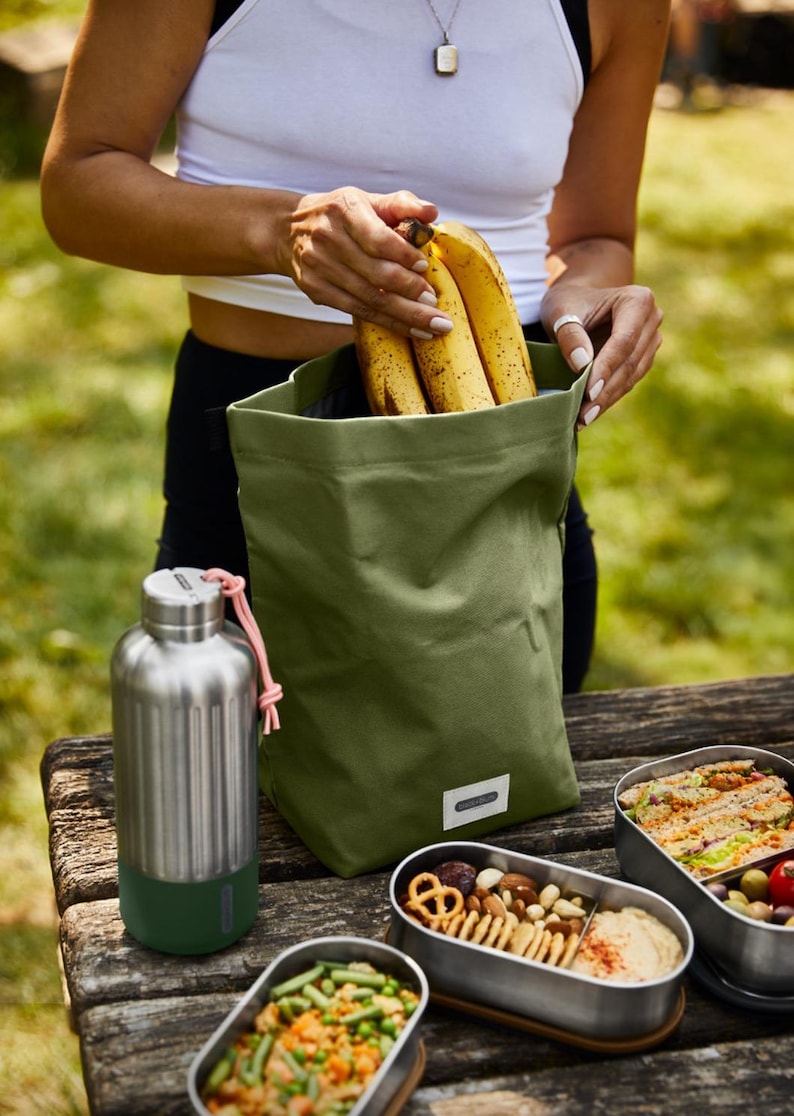May include: A green canvas lunch bag with a pink drawstring, filled with bananas.  A stainless steel water bottle with a green bottom and a silver top is next to the bag.  Three stainless steel lunch containers are on a wooden table, filled with food.