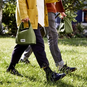 May include: Two people walking on a grassy field, each carrying a green canvas tote bag with a white label. The person on the left is wearing a yellow jacket and black pants, while the person on the right is wearing a red shirt and blue jeans.
