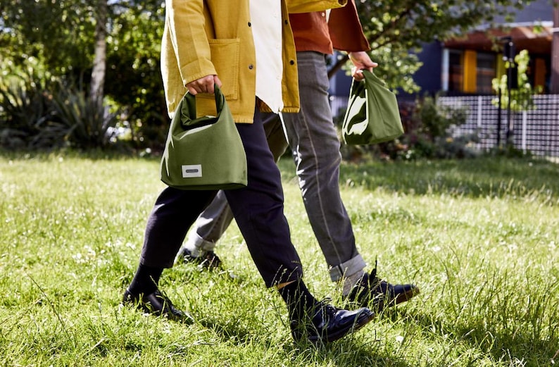 May include: Two people walking on a grassy field, each carrying a green canvas tote bag with a white label. The person on the left is wearing a yellow jacket and black pants, while the person on the right is wearing a red shirt and blue jeans.