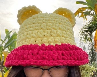 Puede incluir: Un gorro de punto de color amarillo y rosa. El gorro tiene dos solapas en forma de orejas en la parte superior.