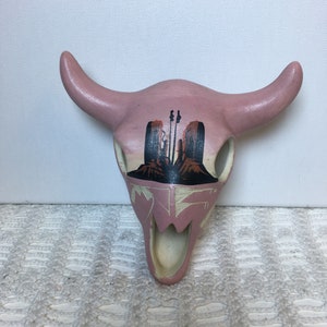 May include: A pink ceramic bull skull with a painted design of a desert landscape with mountains and cacti. The skull has a white interior and two horns.