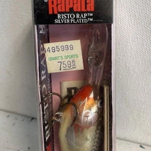 Könnte beinhalten: Ein Rapala Risto Rap 7S Angelköder in einer transparenten Kunststoffbox. Der Köder ist silber, weiß und orange mit drei Haken. Die Verpackung enthält das Rapala-Logo und den Text "Risto Rap" und "Silver Plated".