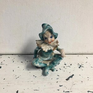 Puede incluir: Figura de cerámica de un elfo sentado, en tonos turquesa y blanco. El elfo lleva un gorro puntiagudo, un cuello con volantes y tiene una expresión traviesa. La figura está sobre una superficie blanca desgastada.