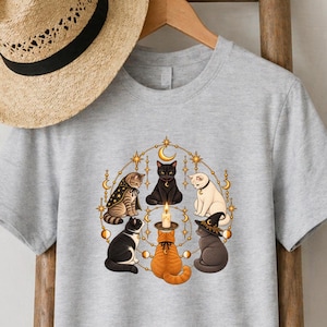 May include: A light grey short-sleeved t-shirt featuring a circular graphic of five cats surrounding a central lit candle. The cats are depicted in various colours including black, white, orange, and tabby, with some wearing small hats and capes.